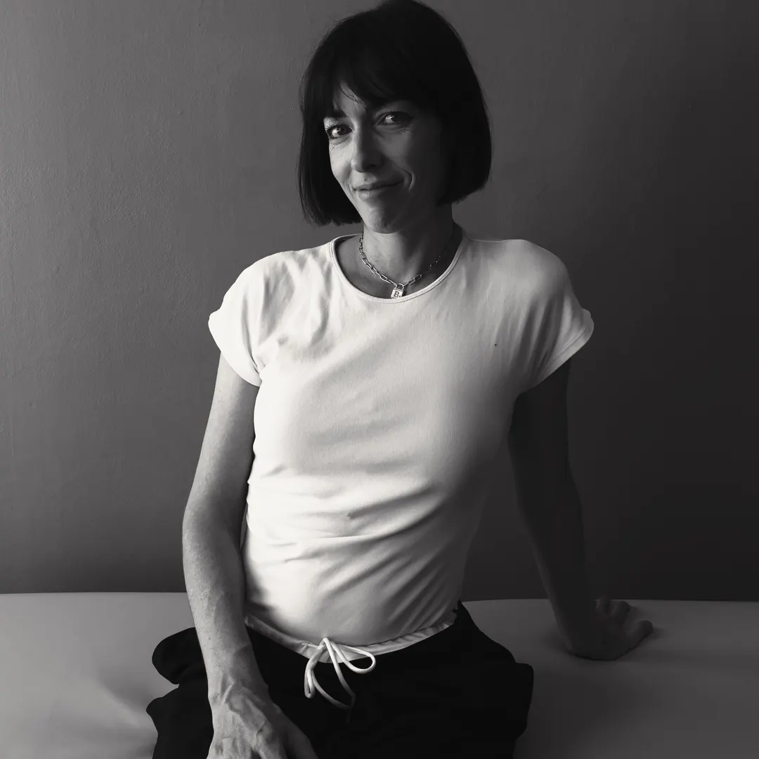 Photographie en noir et blanc de Tiffany Calin assise sur sa table de soin, en tee-shirt blanc et pantalon noir, une main posée derrière elle.